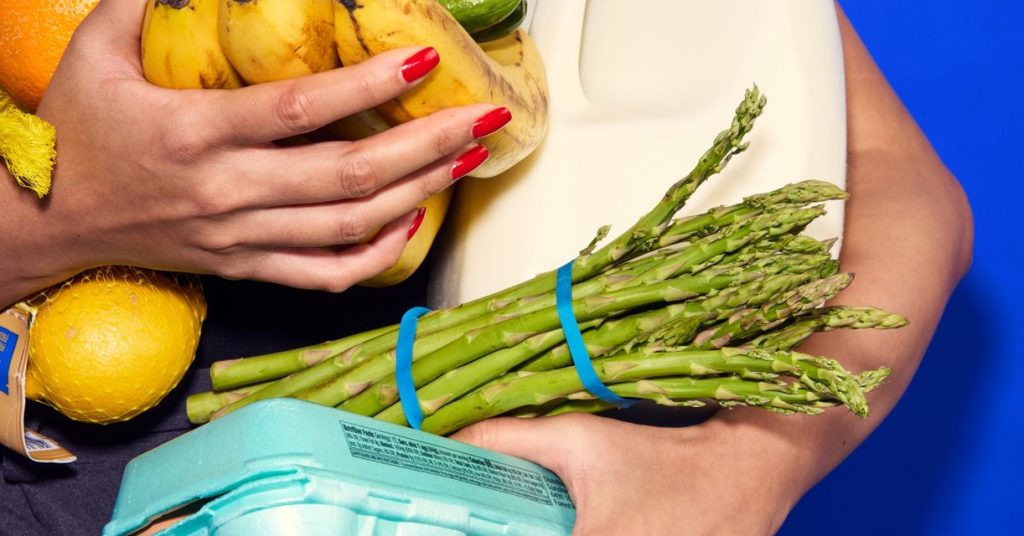 An armful of fresh groceries — bananas, asparagus, eggs, milk and citrus — held against a blue background