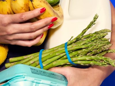 An armful of fresh groceries — bananas, asparagus, eggs, milk and citrus — held against a blue background