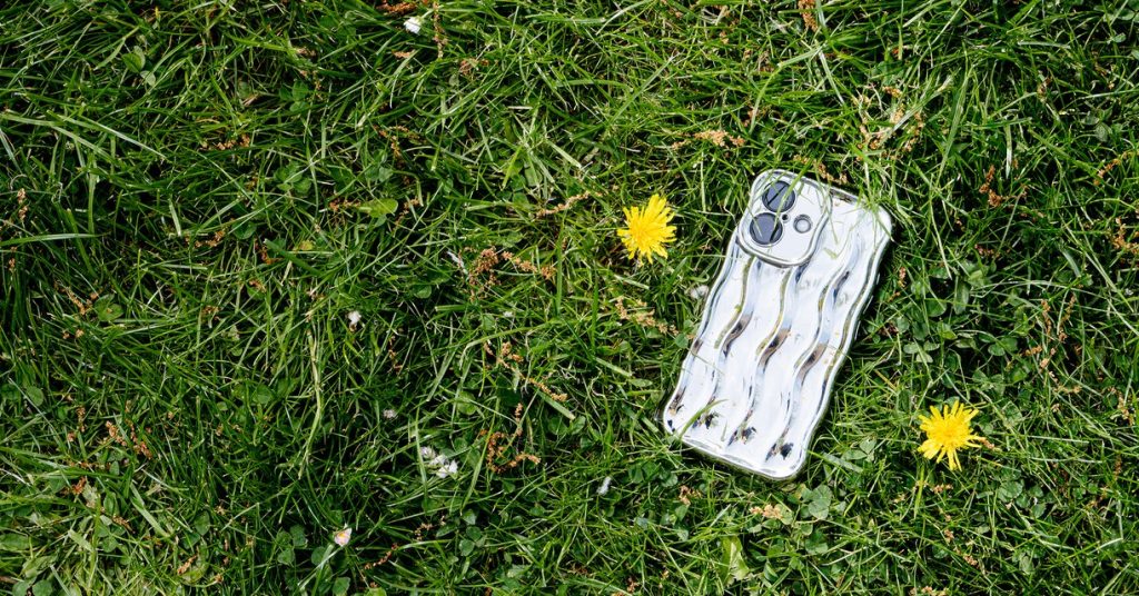 A refurbished iPhone in a clear wavy case lying face down in green grass beside two dandelions