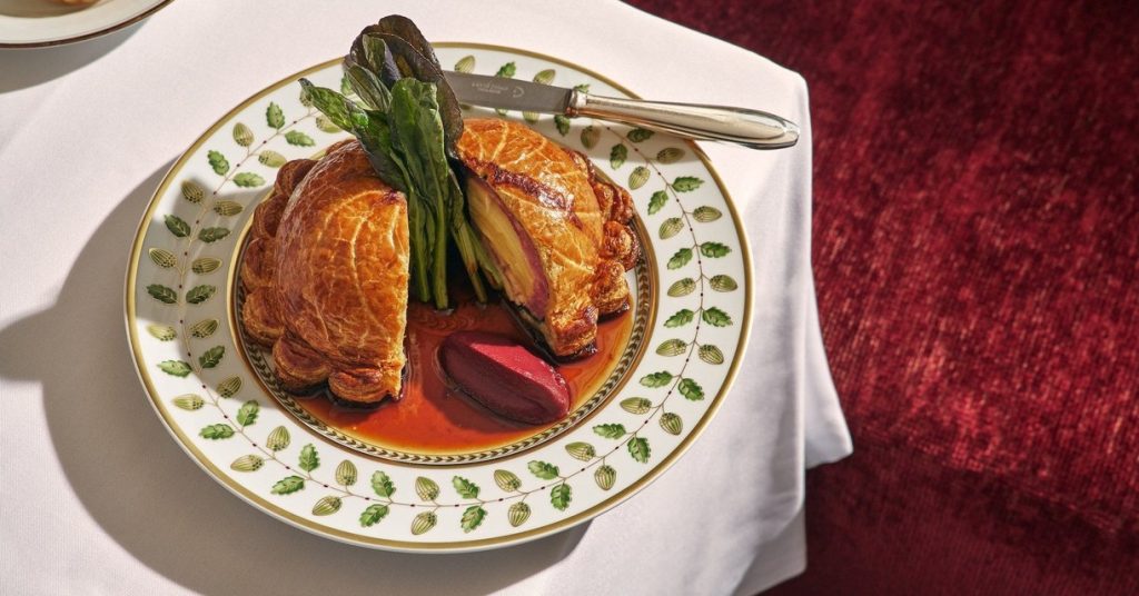A beef Wellington cut in half on a gold-rimmed porcelain plate, set on a white tablecloth beside a red velvet banquette