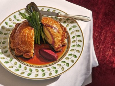 A beef Wellington cut in half on a gold-rimmed porcelain plate, set on a white tablecloth beside a red velvet banquette