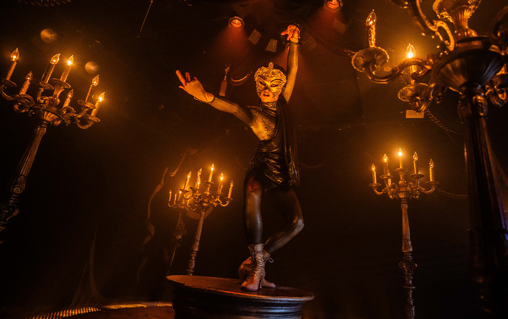 A masked dancer on a small round stage between lit candelabras in Masquerade