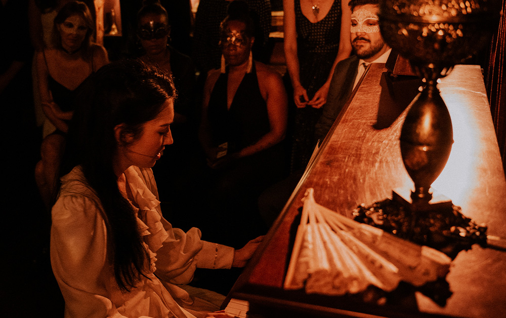 A performer playing piano in Masquerade while masked audience members stand a few feet away