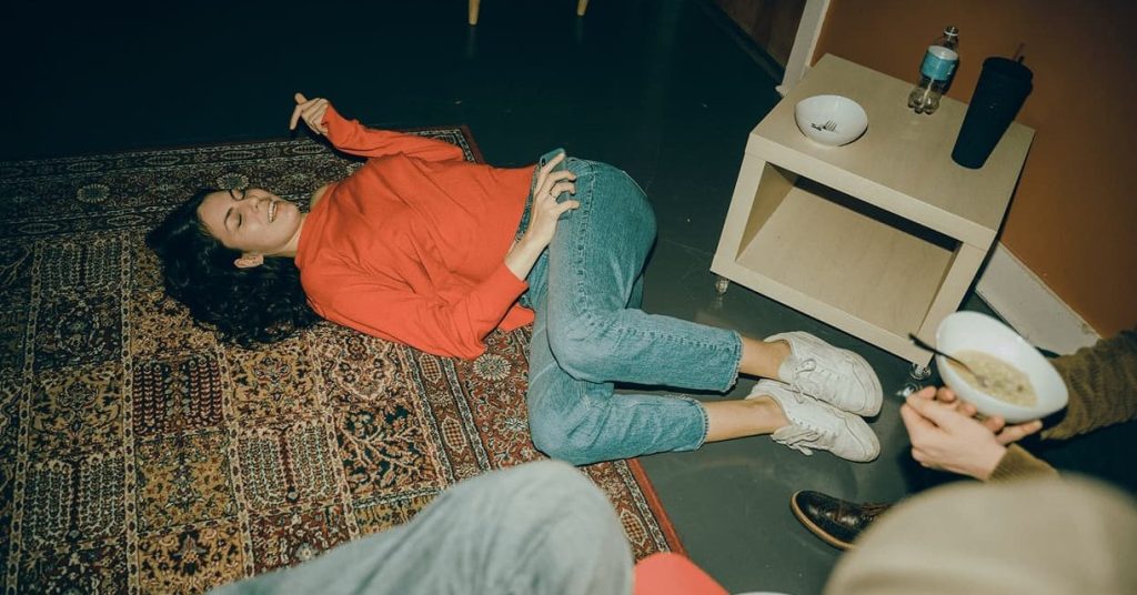 A woman in a red sweater lies laughing on a patterned rug at home, holding her phone
