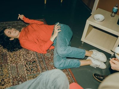 A woman in a red sweater lies laughing on a patterned rug at home, holding her phone