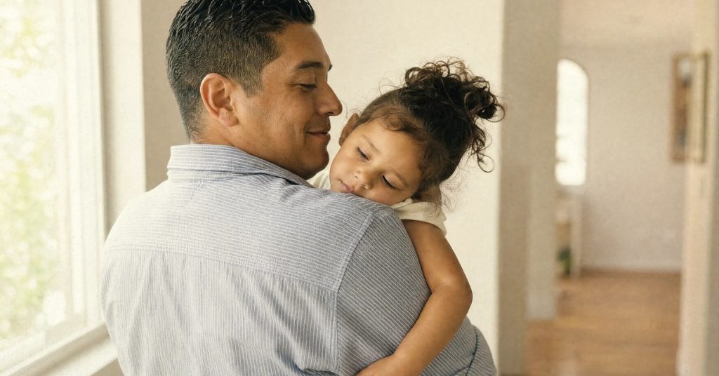 A father holds his sleeping young daughter on his shoulder in a sunlit hallway