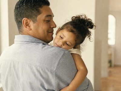 A father holds his sleeping young daughter on his shoulder in a sunlit hallway